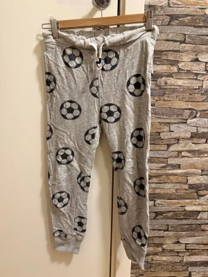 Grå pyjamasbyxor med fotbollsmönster - Mysiga grå pyjamasbyxor med svartvita fotbollar över hela tyget. Byxorna har resår och dragsko i midjan samt muddar vid bensluten. Perfekta för chillkvällar hemma. Materialet känns mjukt och stretchigt.