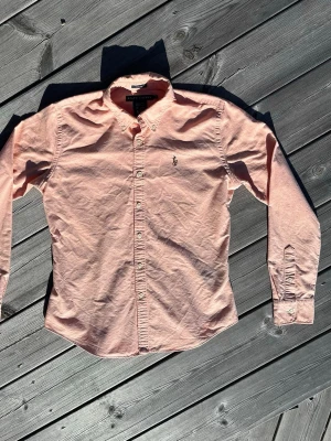 Ljusrosa skjorta från Ralph Lauren - Klassisk ljusrosa skjorta från Ralph Lauren med button-down krage och broderad logga på bröstet. Skjortan har långa ärmar, vita knappar och är tillverkad i mjuk bomull. Perfekt för en clean och stilren look.
