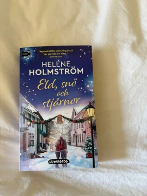 Eld, snö och stjärnor av Holmström - En romantisk och vintrig pocketbok av Heléne Holmström. Omslaget är blått och vitt med snöfall, juldekorerade hus och ett par som kramas på en gata. Perfekt för dig som gillar mysiga julberättelser med kärlek och bakning i centrum.