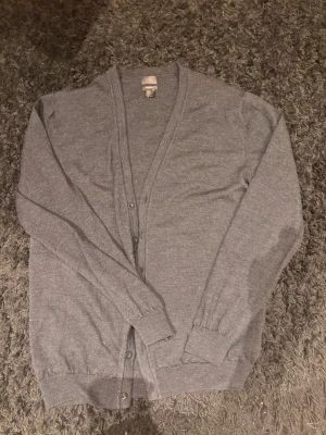 Mörkgrå merinoullskofta från H&M - Stilren mörkgrå kofta från H&M i mjuk 100% merinoull. Klassisk V-ringning, knäppning framtill med sex knappar och ribbade muddar vid ärmslut och nederkant. Perfekt lager på lager-plagg för kyligare dagar eller everyday look. Hör av vid frågor!