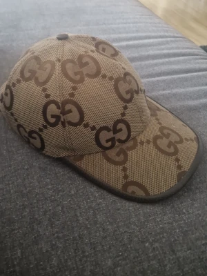 Beige keps från Gucci med GG-mönster - Snygg beige keps från Gucci med det klassiska GG-monogrammet i brunt över hela kepsen. Kepsen har böjd skärm och är tillverkad i ett slitstarkt tygmaterial. Perfekt för dig som vill ha en exklusiv och trendig accessoar. Skriv gärna om ni har frågor svarar direkt
