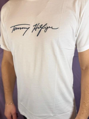 Vit T-shirt från Tommy Hilfiger - Snygg vit T-shirt från Tommy Hilfiger med svart logotyp i skrivstil på bröstet. Klassisk passform och rund halsringning. Tillverkad i mjuk bomull som känns skön mot huden. Perfekt för en clean och stilren look.