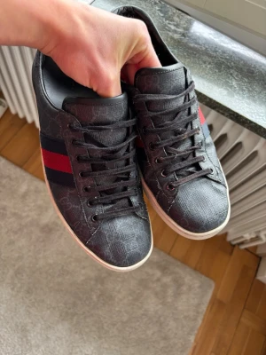 Svarta Gucci ace sneakers - Snygga svarta sneakers från Gucci med klassiskt GG-mönster i material som ser ut som läder. Skorna har en röd och blå rand på sidan och svarta skosnören. Perfekta för dig som vill ha en stilren och lyxig look. Enda defekten bild 4 och lite creasade.