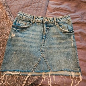 Blå jeanskjol med fransig kant - Säljer en blå jeanskjol i kort modell med slitna detaljer och fransig, rå kant längst ner. Kjolen har klassiska fickor fram och bak, bälteshällor och en cool asymmetrisk söm framtill. Perfekt för dig som gillar en avslappnad och trendig stil.  Kan även hämtas I Partille  E2