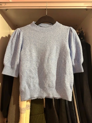 Ljusblå stickad topp från Vero Moda - Ljusblå stickad topp från Vero Moda med puffade korta ärmar och ribbad halsringning. Materialet är mjukt och lätt, perfekt för att skapa en chill och trendig look. Passar dig som gillar en feminin och modern stil.