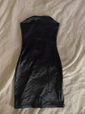 Svart axelbandslös kort klänning - Snygg svart axelbandslös kort klänning i figurnära modell. Klänningen har en slät yta och är tillverkad i ett glansigt material som ger en elegant look. Perfekt för dig som vill ha en enkel men stilren outfit.