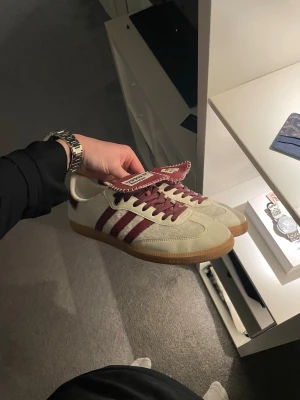 Adidas X Wales Bonner sneakers beige/vinröd - Säljer ett par Adidas Wales Bonner sneakers i beige mocka med vinröda detaljer och snören. Nypris 6999❌ Mitt pris 3499 ✅Skorna har klassiska tre ränder på sidan, gummisula och en unik tungdesign med Wales Bonner-logga. Perfekta för dig som gillar retrostil och snygga detaljer.