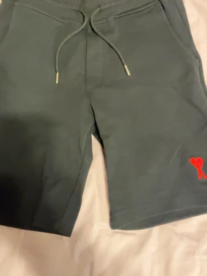 Svarta shorts från Ami Paris - Mörkgröna shorts från Ami Paris med snörning i midjan och två sidofickor. På vänster ben finns Ami Paris ikoniska röda hjärta med bokstaven A broderat. Perfekta för sommaren och tillverkade i mjuk bomull.