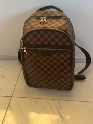 Brun rutig ryggsäck Louis Vuitton - Snygg brun ryggsäck från Louis Vuitton med klassiskt rutigt Damier-mönster. Ryggsäcken har flera fack med dragkedja, justerbara axelband och bärhandtag. Tillverkad i canvas med läderdetaljer och silverfärgade metalldetaljer. Perfekt för dig som gillar lyxig och tidlös stil.