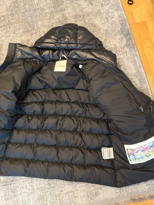 Svart dunjacka från Moncler med huva - Snygg svart dunjacka från Moncler med huva och dragkedja framtill. Jackan har två framfickor med dragkedja och ett stort vitt Moncler-märke på ena ärmen. Klassisk quiltad design och innerficka med dragkedja. Perfekt för kalla vinterdagar.