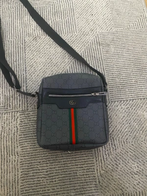 Gucci axelväska med GG-mönster - Snygg axelväska från Gucci i mörkgrått med klassiskt GG-mönster och en grön-röd rand framtill. Väskan har justerbar svart axelrem, dragkedja fram och bak samt detaljer i svart läder. Perfekt för dig som vill ha en stilren och trendig accessoar.