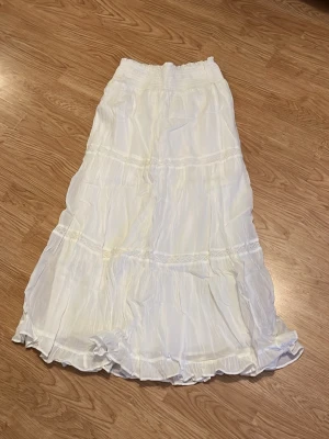 Vit långkjol med volangdetaljer - Luftig vit långkjol med flera volanglager och resår i midjan. Kjolen har en lätt skrynklig struktur och faller snyggt längs benen. Perfekt för en somrig och avslappnad stil.