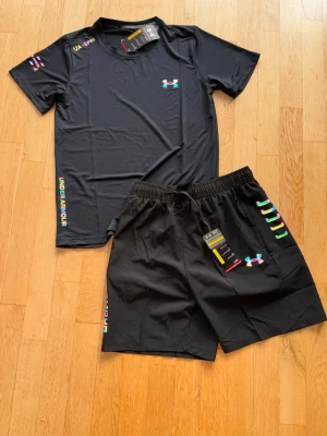 Svart träningsset från Under Armour - Snyggt svart träningsset från Under Armour med t-shirt och shorts. Setet har färgglada loggor och detaljer, samt tryckt Under Armour-text på sidan. Tillverkat i lätt och snabbtorkande material som passar perfekt till träning och sport.