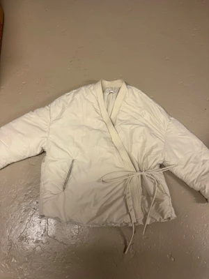 Vit vadderad kimono-inspirerad jacka - Säljer en vit dunjacka i omlottmodell med knytband i midjan. Jackan har två sidofickor och ribbad kant vid halsen. Perfekt för kalla vinterdagar och ger en unik look med sin kimono-inspirerade design. Har en liten blå fläck som kanske går bort i tvätten (tuschpenna) men syns knappt. PRIS KAN ALLTID DISKUTERAS! 