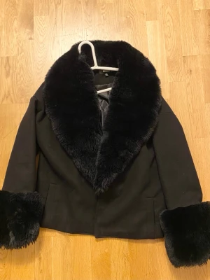 Svart pälsjacka med bred krage - Snygg svart pälsjacka med bred, fluffig fuskpäls runt kragen och ärmsluten. Jackan har en rak passform och två fickor framtill. Perfekt för dig som vill ha en statementjacka med lyxig känsla och cool attityd.