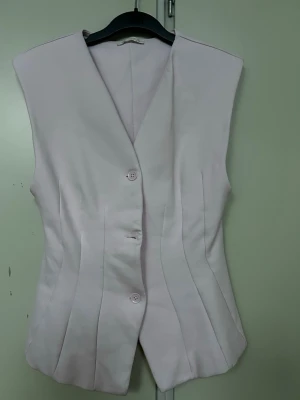 Ljuslila figursydd väst med knappar - Säljer en ljuslila väst med figurnära passform och markerade sömmar. Västen har V-ringning och stängs med tre matchande knappar framtill. Materialet känns mjukt och har en lätt struktur. Perfekt för att lyfta din outfit med en stilren touch.