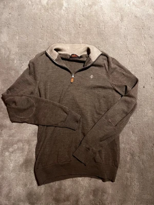 Brun merinoullströja Morris - Snygg brun tröja från Morris i 100% merinoull med en quarter zip och beige ribbad krage. Tröjan har diskret logga broderad på bröstet och förstärkta armbågar. Perfekt för lager på lager och stilrena outfits.