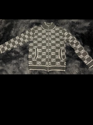 Svart stickad ziptröja från Gucci - Säljer en svart stickad ziptröja från Gucci med vit GG-monogram över hela plagget. Tröjan har vita detaljer vid muddar, krage och fickor samt två framfickor och dragkedja framtill. Perfekt för dig som gillar lyxiga streetwear-vibbar.