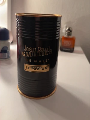 Jean Paul Gaultier Le Male Le Parfum - Jean Paul Gaultier Le Male Le Parfum i en elegant svart och guld metallburk. Flaskan har en unik design formad som en manlig torso i svart glas med gulddetaljer och står på en röd sammetsliknande bas. En ikonisk och lyxig herrdoft. Parfymen är nästintill oanvänd taget 2-3 sprut av den!
