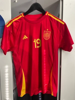 Spaniens matchtröja Lamine Yamal #19 - Röd matchtröja från Adidas för Spaniens landslag med gula detaljer, nummer 19 och Lamine Yamal på ryggen. Tröjan har korta ärmar, rund hals och gula ränder på axlarna. Tillverkad i lätt och ventilerande funktionsmaterial, perfekt för fotboll. Helt ny använt den Max 5 gånger 