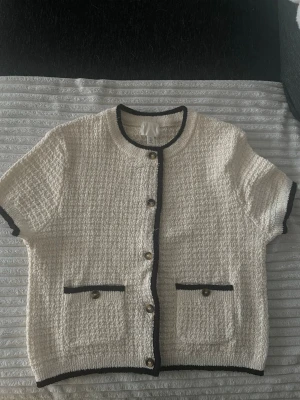 Vit kortärmad kofta med svarta detaljer - Stilren vit kofta i bouclé-liknande material med korta ärmar och svarta kontrastkanter. Rund halsringning, knappar framtill och två fickor med knappdetaljer. Perfekt för en chic och trendig look.🤗 Aldrig använd, endast testad💕Priset kan diskuteras vid snabb affär😊