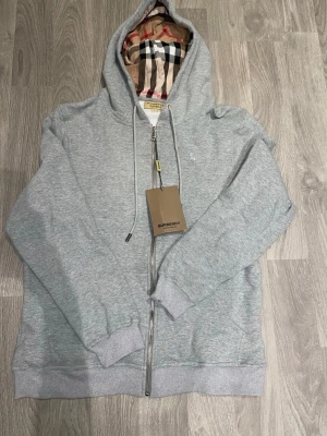 Grå zip hoodie från Burberry - Snygg ljusgrå hoodie från Burberry med dragkedja framtill och klassiskt rutigt foder i huvan. Tillverkad i mjuk bomullsblandning, har ribbade muddar och justerbar dragsko i huvan. Perfekt för en avslappnad och stilren look. ❗️OBS ❗️- Skriv till mig innan köp! 🙌🏼