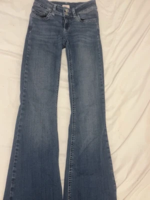 Blå bootcut jeans från Nelly - Snygga blå bootcut jeans från Nelly med dubbla knappar i midjan och klassisk femficksdesign. Jeansen har en ljus tvätt och markerade sömmar som ger en trendig look. Perfekta för dig som gillar en classy stil.