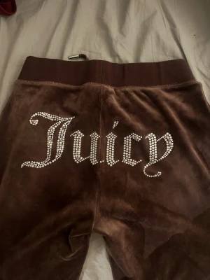 Bruna Juicy Couture mjukisbyxor - Mörkbruna mjukisbyxor i velour från Juicy Couture med strassdetaljer och stor text 'Juicy' på baksidan. Byxorna har bred resår i midjan, dragsko och fickor. Perfekta för en chill och trendig look.