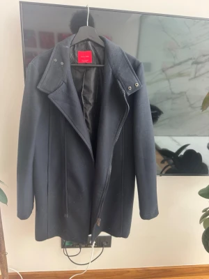 Mörkblå kappa från Zara - Stilren mörkblå kappa från Zara med bred krage och asymmetrisk dragkedja. Kappan har en klassisk rak passform och är helfodrad för extra komfort. Perfekt för dig som vill ha en tidlös och snygg look. Materialet är troligtvis ullblandning.