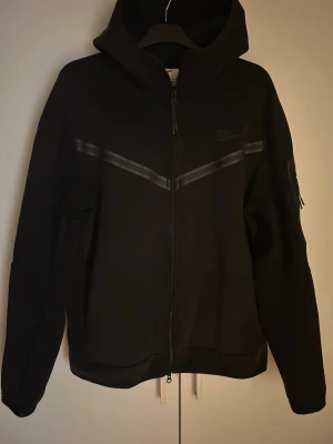 Nike Tech fleece - Säljer en svart zip-hoodie från Nike. Stilren och bekväm modell med huva, dragkedja och diskreta reflexdetaljer. Fint skick utan synliga defekter. Lite solblekt färg men annars i ett gott skick, har kommit till användning. Pris kan diskuteras vid snabb affär! Hör av er vid frågor eller funderingar🤩👌🏼