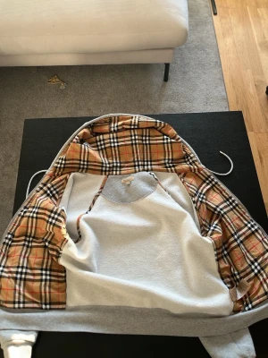 Grå hoodie med Burberry-mönstrat foder - Snygg grå hoodie med klassiskt Burberry-rutigt foder i huvan och insidan. Hoodien har dragsko i huvan och ribbade muddar. Perfekt för dig som vill ha en stilren look med en twist av lyxigt mönster. Materialet är mjukt och bekvämt. Passar alldelses utmärkt för er som har storlek S 