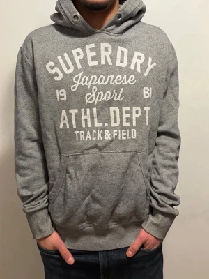 Extremt sällsynt Grå Superdry hoodie med tryck - Snygg grå hoodie från Superdry med stort vitt tryck på bröstet där det står 'Superdry Japanese Sport ATHL. DEPT Track & Field'. Hoodien har huva med dragsko och en klassisk magficka. Perfekt för en avslappnad och sportig stil.