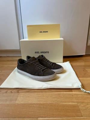  Axel Arigato skor - Mörkbruna sneakers i mocka från Axel Arigato med klassisk låg siluett och vita gummisulor. Allt OG ingår - Låda, Dustbag, Omslagspapper, Klistermärken. Skorna är i toppenskick och har inga defekter eller skador (finns tecken på användning på sulan) Skick 8,5/10                           Pris 1099kr - Pris kan diskuteras                                                               Tveka inte vid frågor eller funderingar