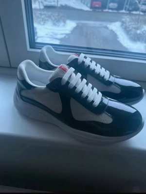 Svarta och vita prada cups prada skor  - Snygga sneakers från Prada med svart och vit färgkombination. Ovandelen är i mesh och glansigt läder, med vita skosnören och en röd Prada-logga på plösen. Klassisk låg modell som passar till streetwear och casual outfits.