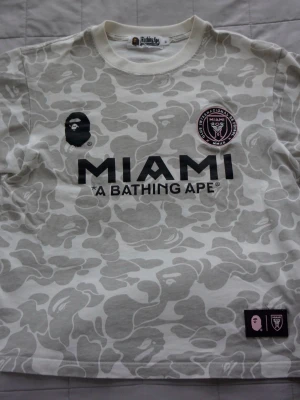 BAPE x Inter Miami | Camo t-shirt - Snygg vit och grå camo t-shirt från BAPE i samarbete med Inter Miami. Stor logga på bröstet med texten 'MIAMI' och 'A BATHING APE', samt klubbmärke och BAPE-ikon. Tillverkad i mjuk bomull och har klassisk passform med korta ärmar. Tröjan är väldigt limited och därför extremt eftertraktad! 