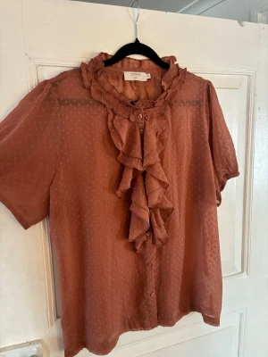 Rostbrun volangblus från Cream - Söt rostbrun blus från Cream med korta ärmar, volangdetaljer längs knäppningen och krage samt diskret prickigt mönster i tyget. Blusen är tillverkad i ett lätt, transparent material som ger en luftig känsla.