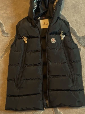 Svart dunväst  med huva - Snygg svart dunväst med vita logotexter längs huvkanten. Västen har två dragkedjeförsedda fickor framtill och klassisk Moncler-logga på bröstet. Tillverkad i glansigt material och fodrad med dun för extra värme. 