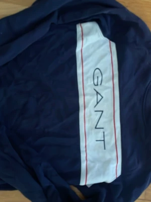 Marinblå hoodie från GANT - Snygg marinblå hoodie från GANT med stort vitt fält och röda linjer över bröstet där GANT-loggan är tryckt. Klassisk passform med långa ärmar och ribbade muddar. Perfekt för dig som gillar stilrena och sportiga plagg.