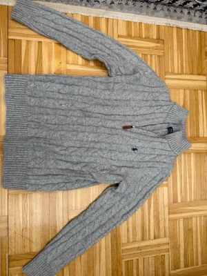 Grå kabelstickad tröja Polo Ralph Lauren - Snygg grå kabelstickad tröja från Polo Ralph Lauren med halv dragkedja och ribbade muddar. Tröjan har det klassiska broderade logotypen på bröstet och är gjord i mjukt ullmaterial. Perfekt för lager på lager och en stilren look.