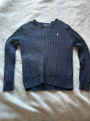 Mörkblå kabelstickad tröja Polo Ralph Lauren - Kabelstickad mörkblå tröja från Polo Ralph Lauren med rund halsringning och vit broderad logga på bröstet. Tröjan har långa ärmar och ribbade muddar vid ärmslut och nederkant. Perfekt för lager på lager och klassisk stil.