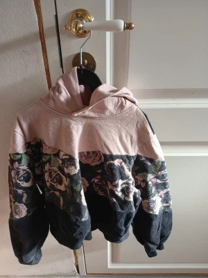 Blommig hoodie i rosa och svart - Snygg hoodie med huva i en mix av ljusrosa och svart. Framsidan har ett blommigt mönster i dova färger som ger en cool kontrast till den enfärgade huvan och nederdelen. Perfekt för dig som gillar unika plagg med lite edge. Mjuk bomullskänsla och avslappnad passform.