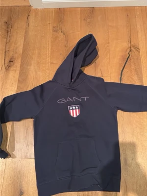 Mörkblå hoodie från GANT med logga - Snygg mörkblå hoodie från GANT med stor huva och känguruficka. På bröstet finns GANTs klassiska logga och ett broderat emblem med stjärnor och rödvita ränder. Perfekt för en avslappnad och sportig stil.