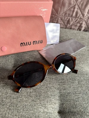 Bruna ovala solglasögon från Miu Miu - Säljer ett par snygga ovala solglasögon från Miu Miu med brunmelerad båge och mörka glas. Kommer med originalfodral och certifikat. Perfekt accessoar för dig som vill ha en trendig och unik look.