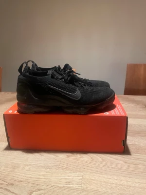 Svarta Nike Air VaporMax sneakers - Säljer ett par helsvarta Nike Air VaporMax sneakers med stickad ovandel och klassisk Swoosh på sidan. Skorna har en chunky sula med synliga luftbubblor och snörning framtill. Perfekta för dig som gillar sportig och clean stil.