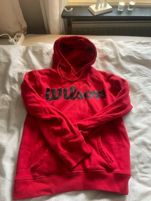 Röd hoodie från Wilson med logga - Snygg röd hoodie från Wilson med stor svart logga på bröstet. Hoodien har huva med dragsko och en klassisk känguruficka framtill. Tillverkad i mjukt och bekvämt material som passar perfekt för chill eller sportiga dagar.