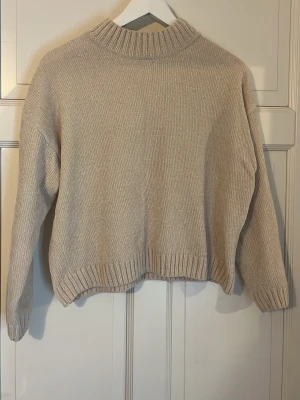 Beige stickad tröja - En stilren beige stickad tröja med ribbad krage och ribbade muddar vid ärmslut och nederkant. Tröjan har en avslappnad passform och är gjord i ett mjukt, tjockt material som passar perfekt för kyligare dagar.