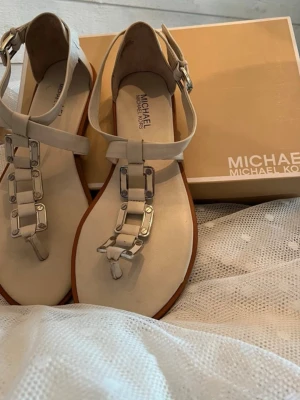 Beige sandaler från Michael Kors - Stilrena beige sandaler från Michael Kors med T-rem och silverfärgade kedjedetaljer framtill. Sandalerna har justerbart spänne vid ankeln och en platt sula i brunt. Tillverkade i läder med metalliska detaljer för en trendig look. Storlek 9M. Använda tre gånger.