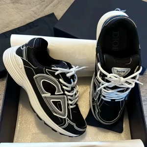 Svarta Dior sneakers med vita detaljer - Snygga svarta Dior sneakers med vita och grå detaljer samt vita skosnören. Skorna har en chunky sula och en modern design med kontrasterande paneler och logga på plösen. Perfekta för dig som vill ha en exklusiv och trendig look.