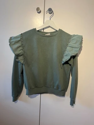 Grön sweatshirt med volang från Kappahl - Snygg grön tröja från Kappahl med volangdetaljer på axlarna. Tröjan har rund halsring, långa ärmar och ribbade muddar. Perfekt för dig som vill ha en trendig och bekväm look.