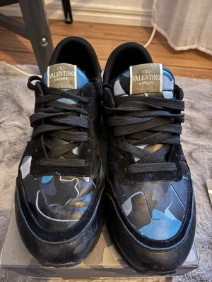 Valentino Garavani camo sneakers - Snygga sneakers från Valentino Garavani med svart bas och blått, grått och vitt camomönster. Skorna har svarta skosnören och en klassisk låg silhuett. Märkeslogga på plösen och cool design som sticker ut. Perfekta för dig som vill ha något unikt och trendigt. Med extra snören påse och låda